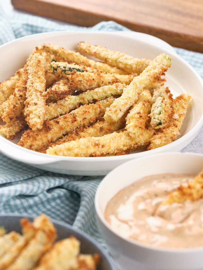 A white bowl filled with crispy, breaded zucchini fries next to a small bowl of dipping sauce.