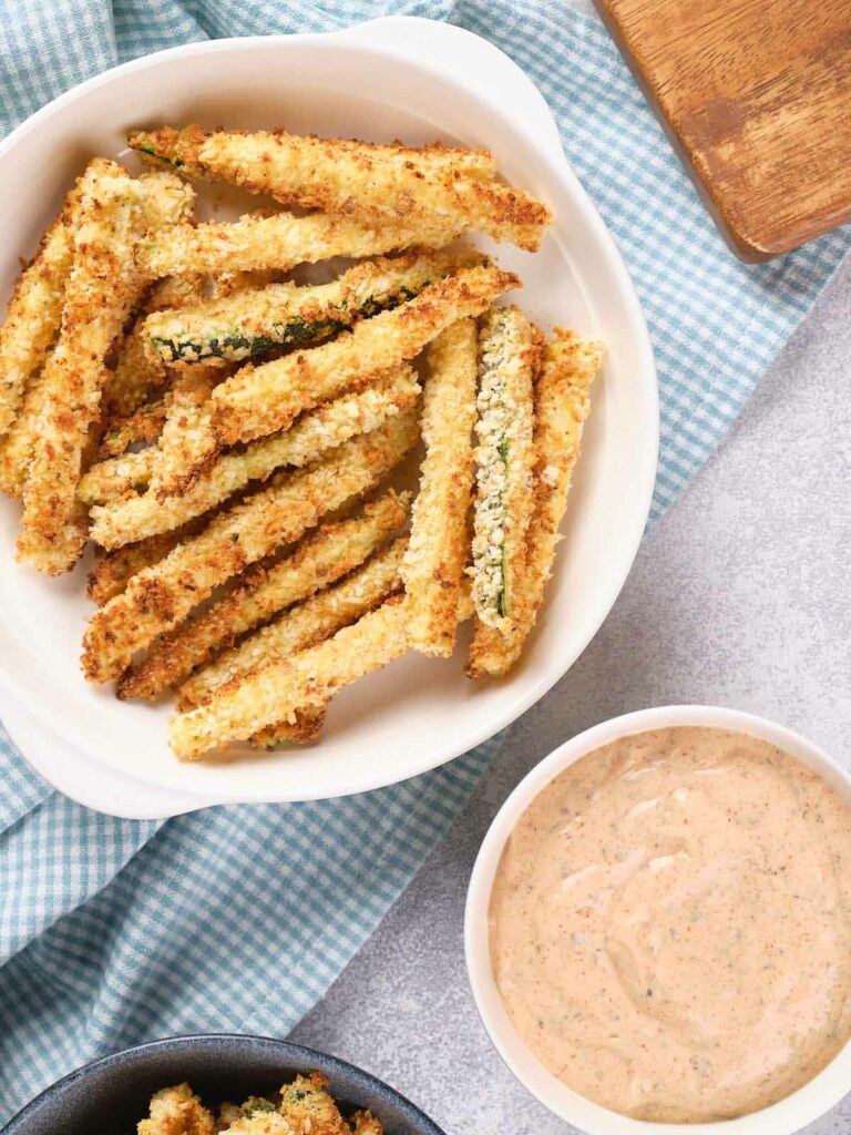 A white bowl filled with crispy breaded zucchini fries next to a bowl of dipping sauce, placed on a blue and white checkered cloth.