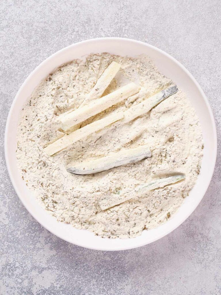 A white bowl containing zucchini sticks coated in a light flour mixture on a textured grey surface.