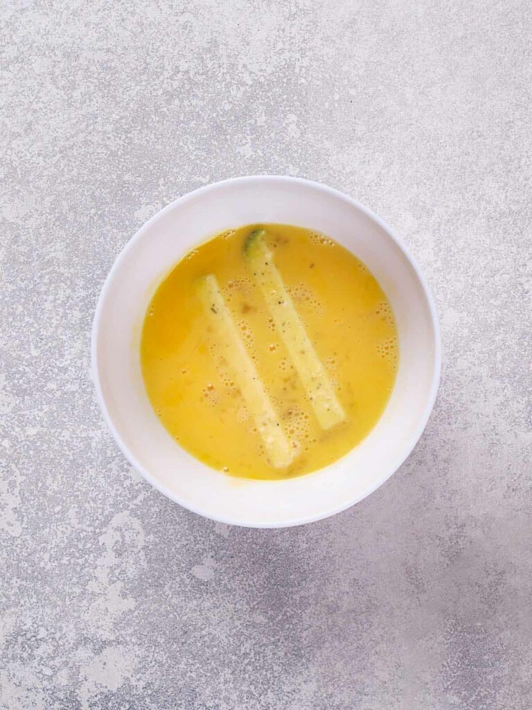 A white bowl containing beaten yellow eggs with two zucchini sticks submerged in the center.