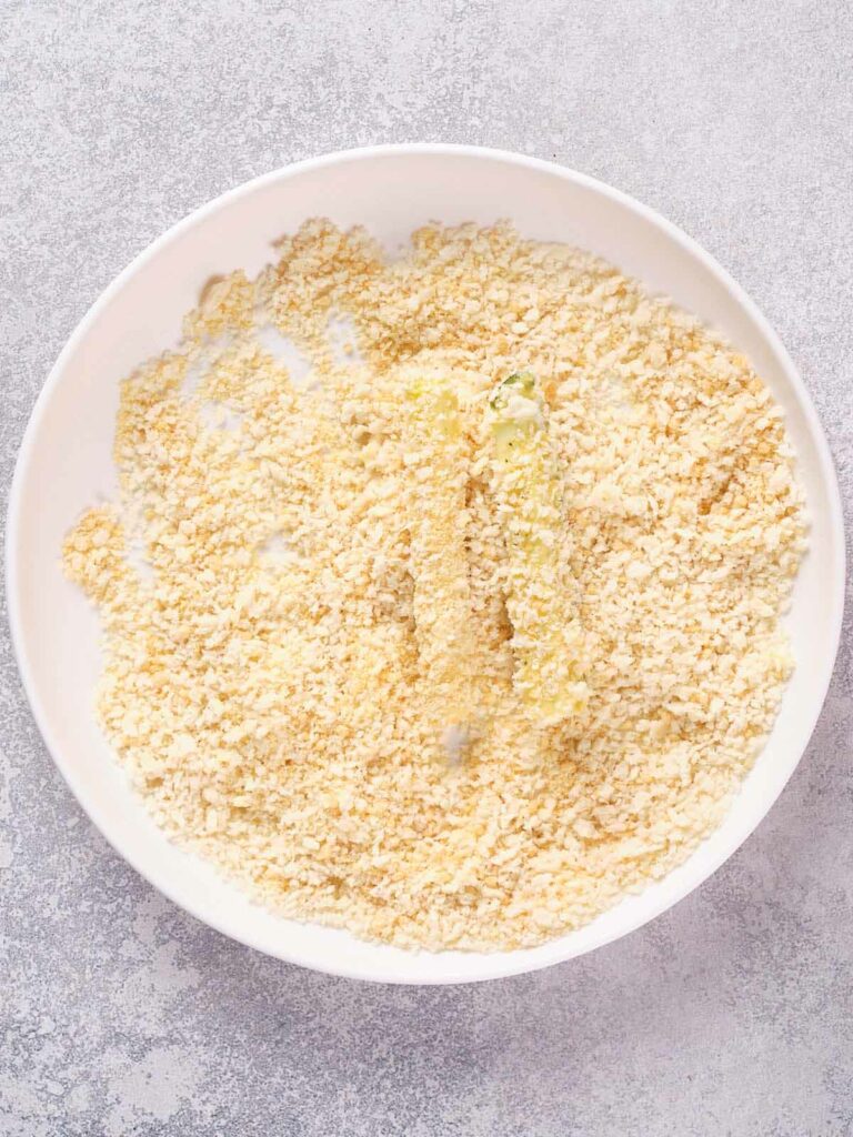 A bowl with two breaded zucchini sticks lying on a bed of breadcrumbs.