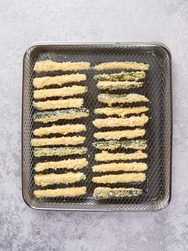 A wire mesh tray holding breaded zucchini fries arranged in parallel rows on a light-colored surface.