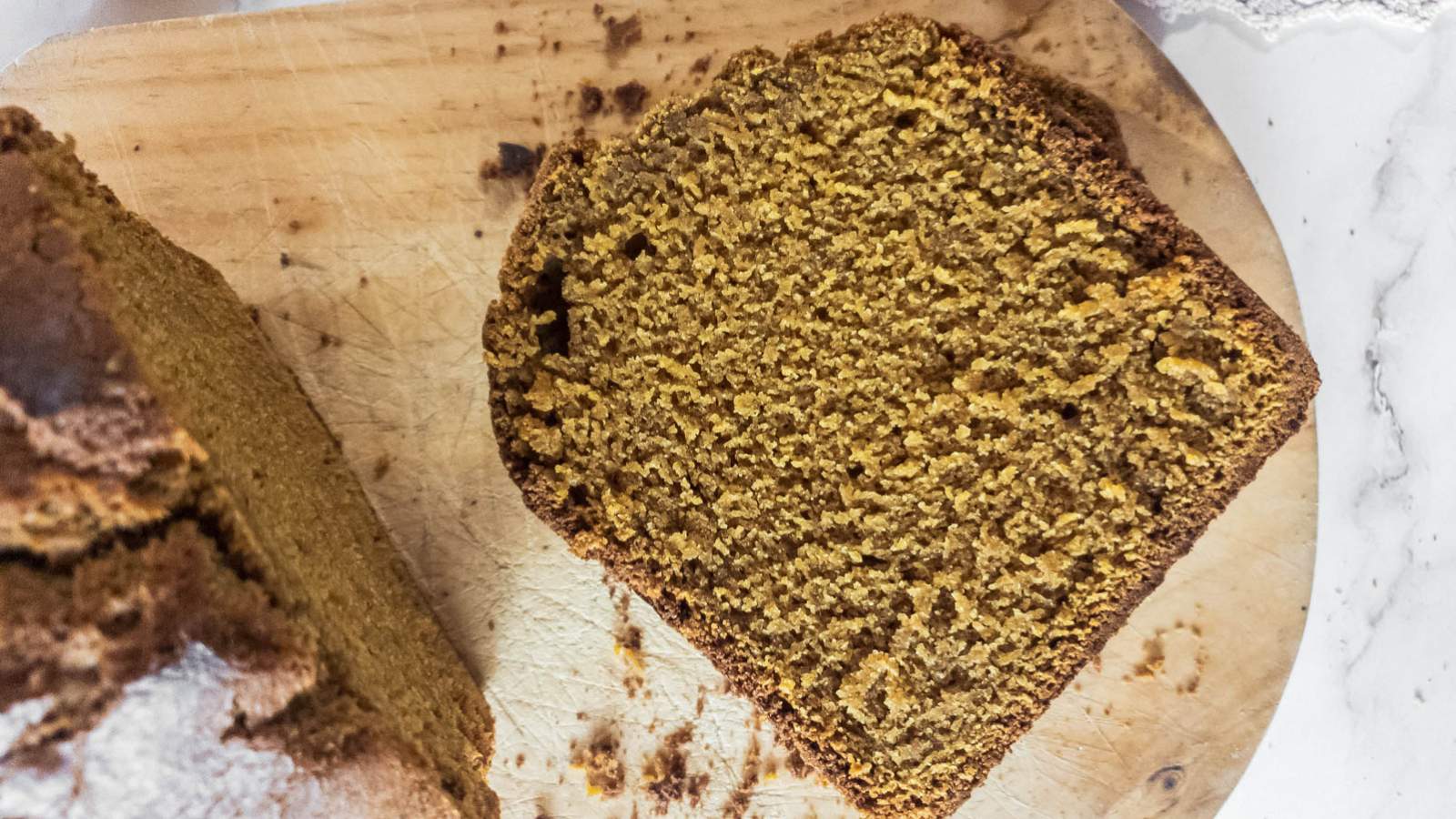 A slice of brown bread rests on a wooden cutting board next to the main loaf.