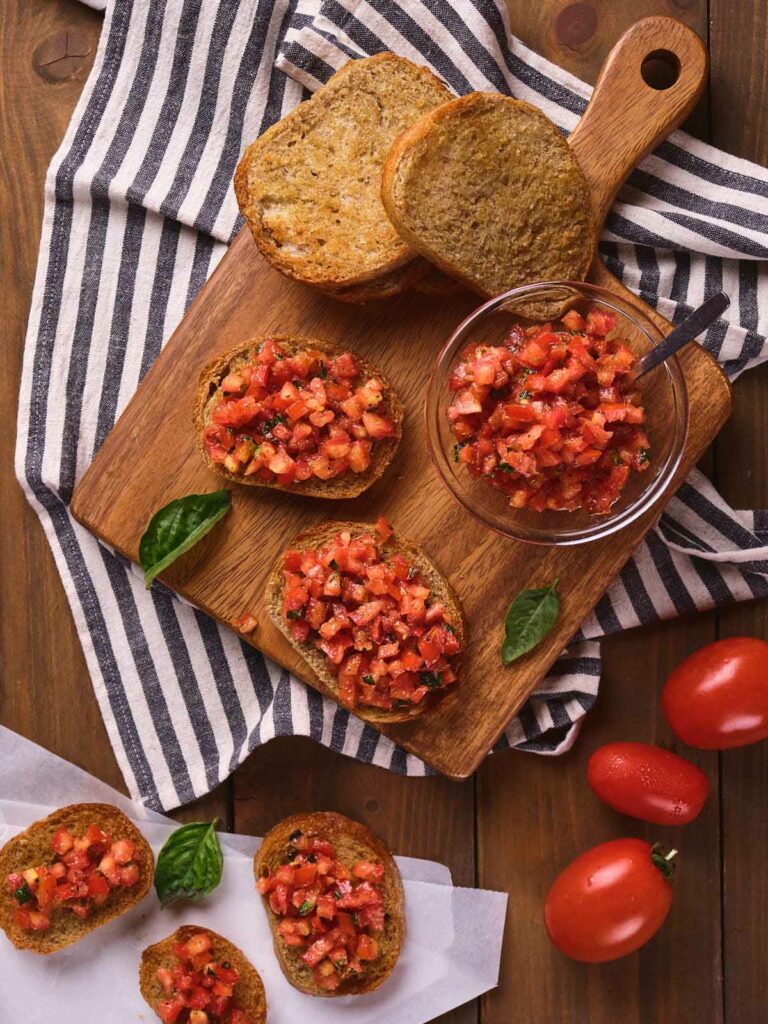 A wooden cutting board with bruschetta topped with diced tomatoes and herbs.