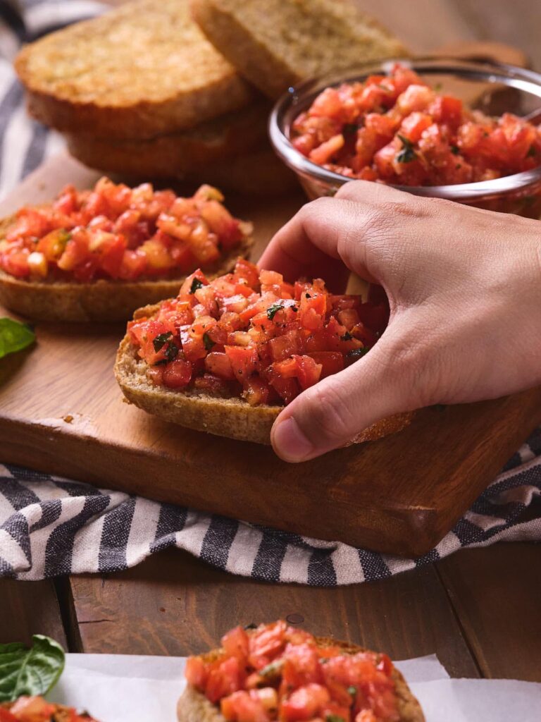 A hand holding a slice of bruschetta al pomodoro on a wooden board in the background.