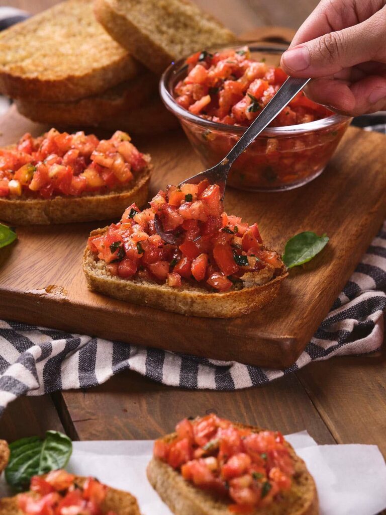 A hand using a spoon to place tomato bruschetta mixture onto a piece of toasted bread on a wooden board.