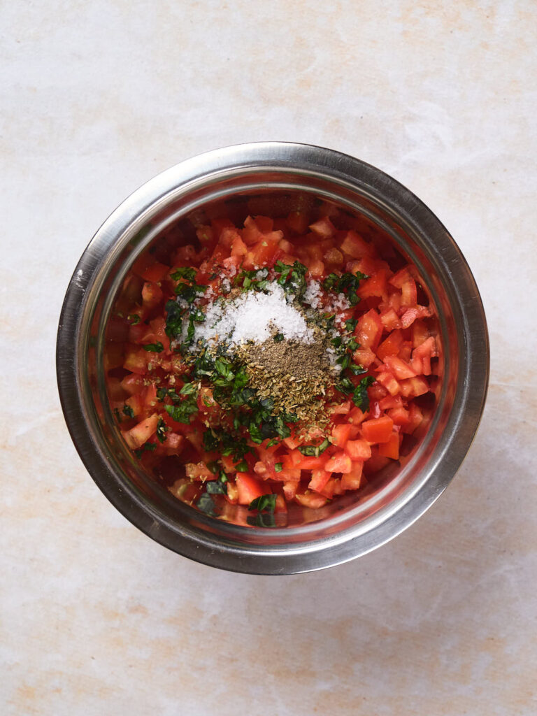 A stainless steel bowl contains diced tomatoes, chopped herbs, and seasonings on a light-colored countertop.