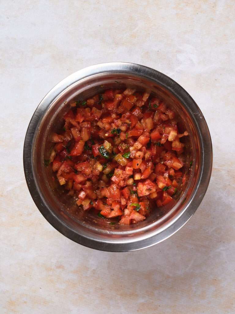 A stainless steel bowl containing diced tomatoes and herbs, placed on a light countertop.