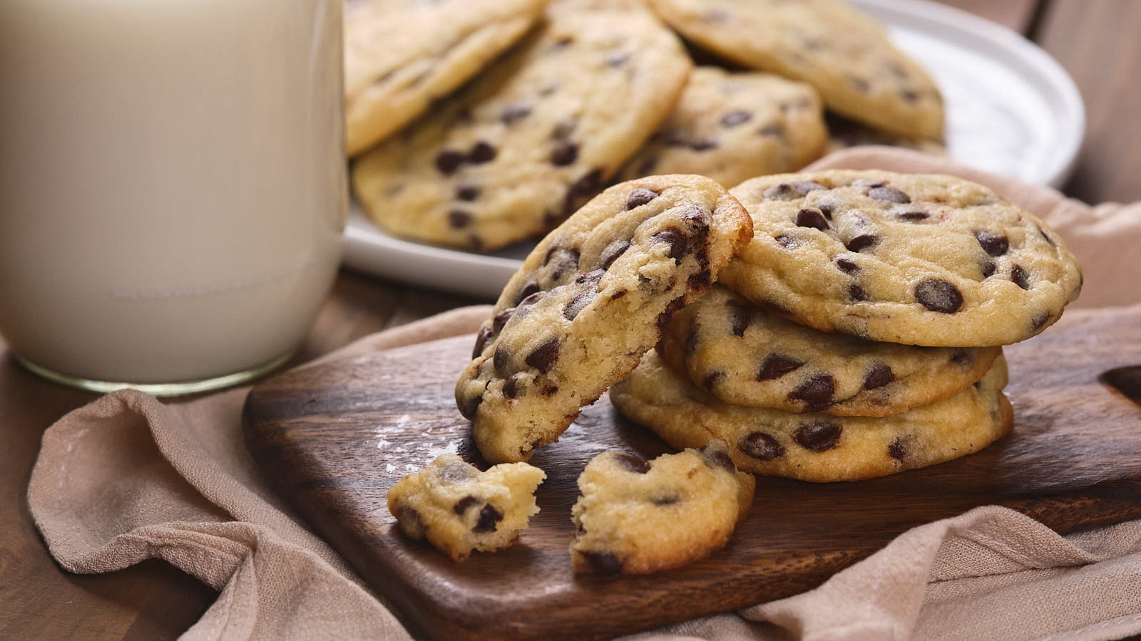 A stack of chocolate chip cookies on a wooden board with a glass of milk nearby. 