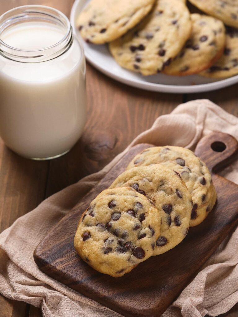 A wooden board with three chocolate chip cookies, a jar of milk, and a plate of more cookies in the background.