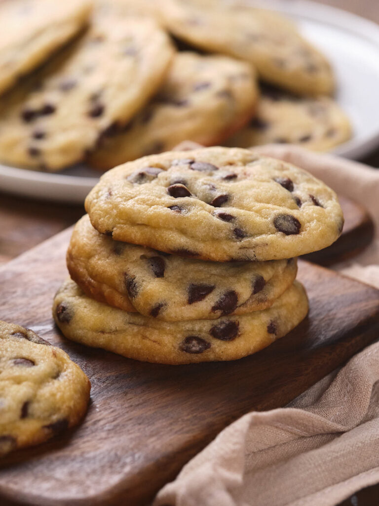 A stack of chocolate chip cookies is placed on a wooden board, with more cookies on a plate in the background.