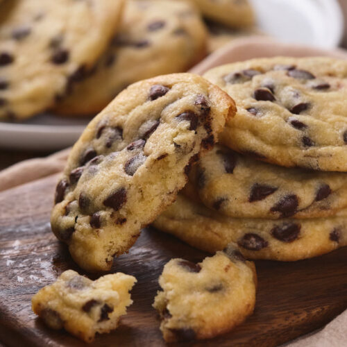 A stack of chocolate chip cookies, with one partially eaten cookie in front of them, placed on a wooden cutting board.