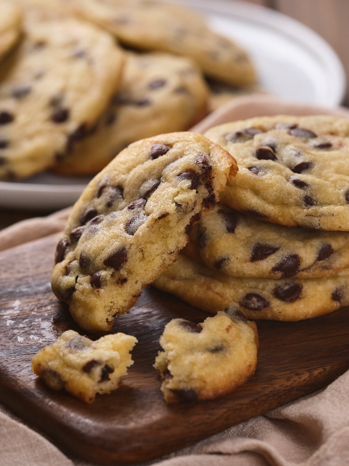 A stack of chocolate chip cookies, with one partially eaten cookie in front of them, placed on a wooden cutting board.