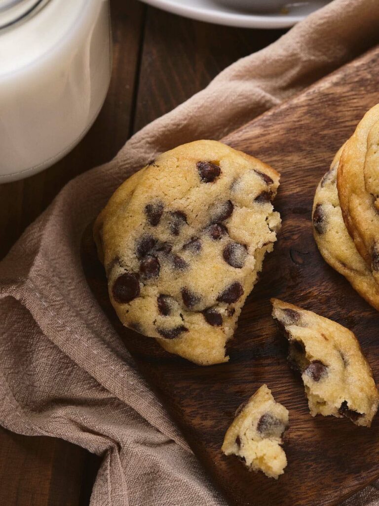 A chocolate chip cookie, partially broken, on a wooden surface with a glass of milk nearby.