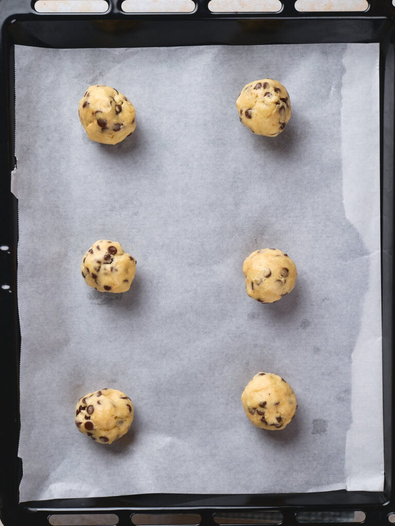 Six balls of chocolate chip cookie dough placed evenly on a parchment-lined baking tray.