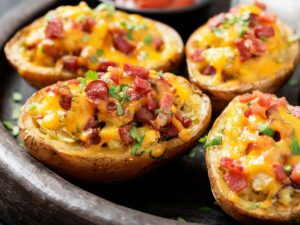 A close-up of four baked potato halves topped with melted cheese, diced bacon, and chopped parsley, served on a dark plate.