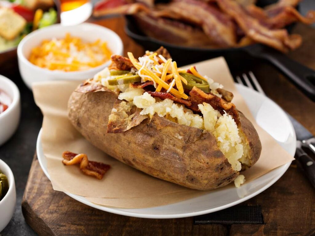 A baked potato topped with bacon bits, shredded cheese, and sliced green peppers on a white plate.