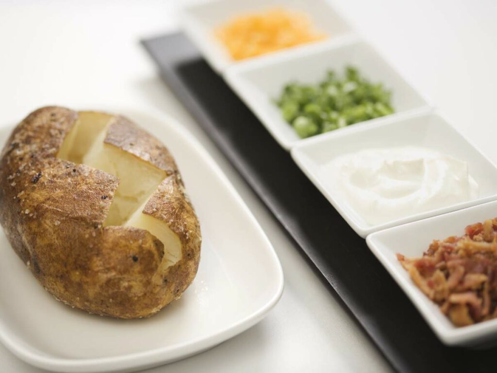 A baked potato with a cross-cut sits on a white plate beside a tray of toppings.