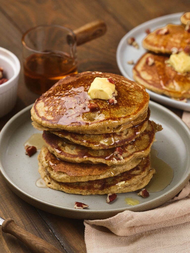 A stack of pumpkin pancakes topped with a pat of melting butter and chopped nuts on a gray plate.