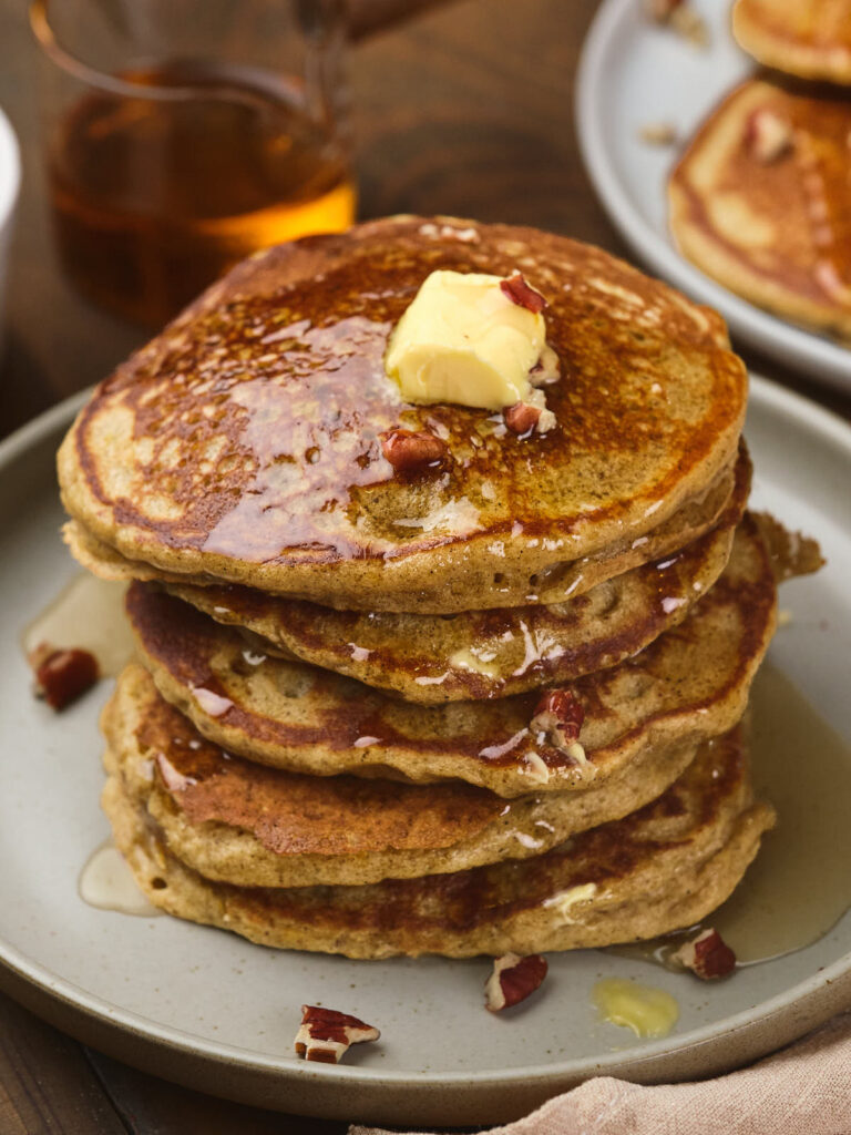 A stack of pumpkin pancakes topped with butter, syrup, and small chopped nuts.