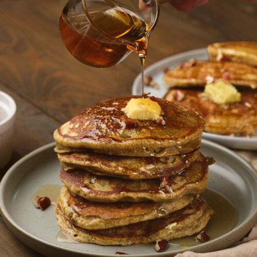 A hand pouring syrup onto a stack of pumpkin pancakes topped with a pat of butter, with more pancakes in the background on a wooden table.