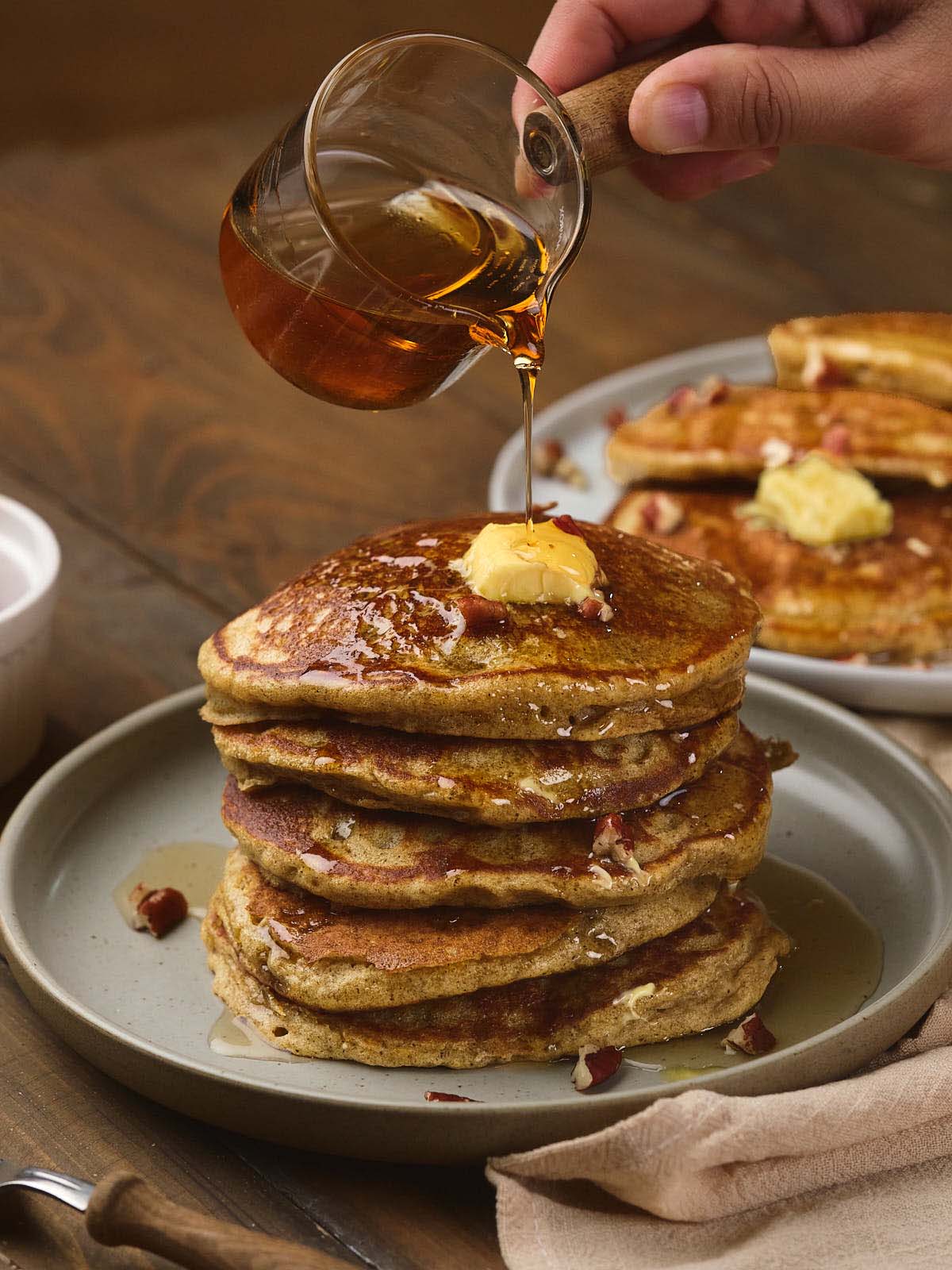 A hand pouring syrup onto a stack of pumpkin pancakes topped with a pat of butter, with more pancakes in the background on a wooden table.