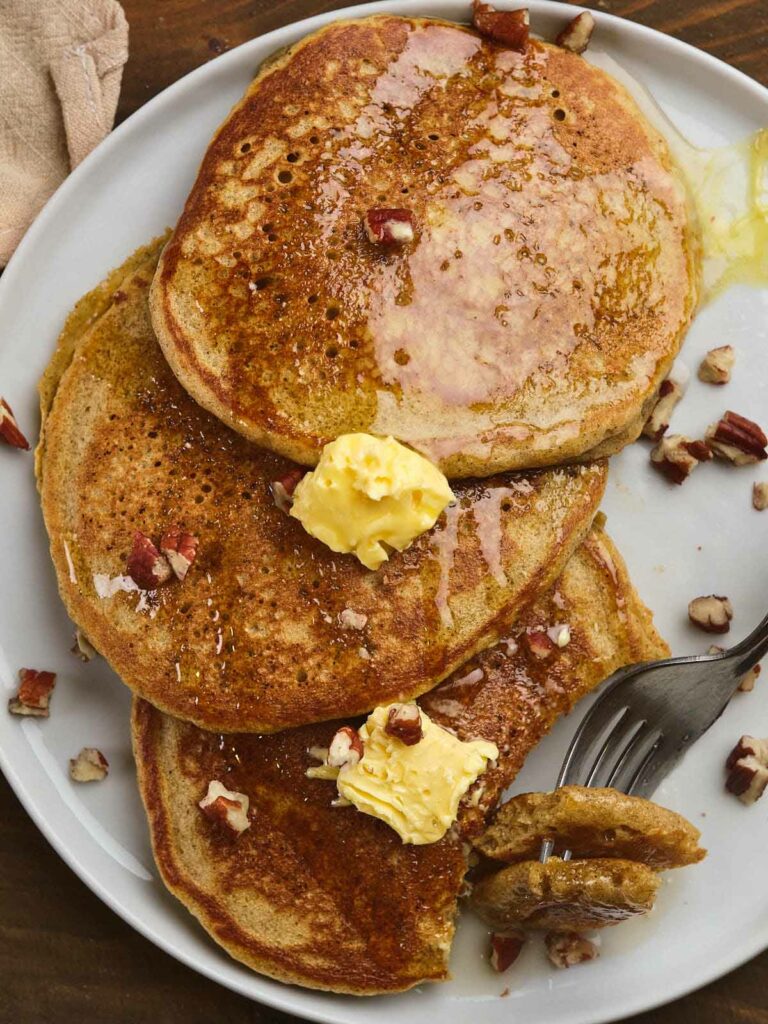 A plate of three pumpkin pancakes topped with butter, syrup, and chopped nuts.