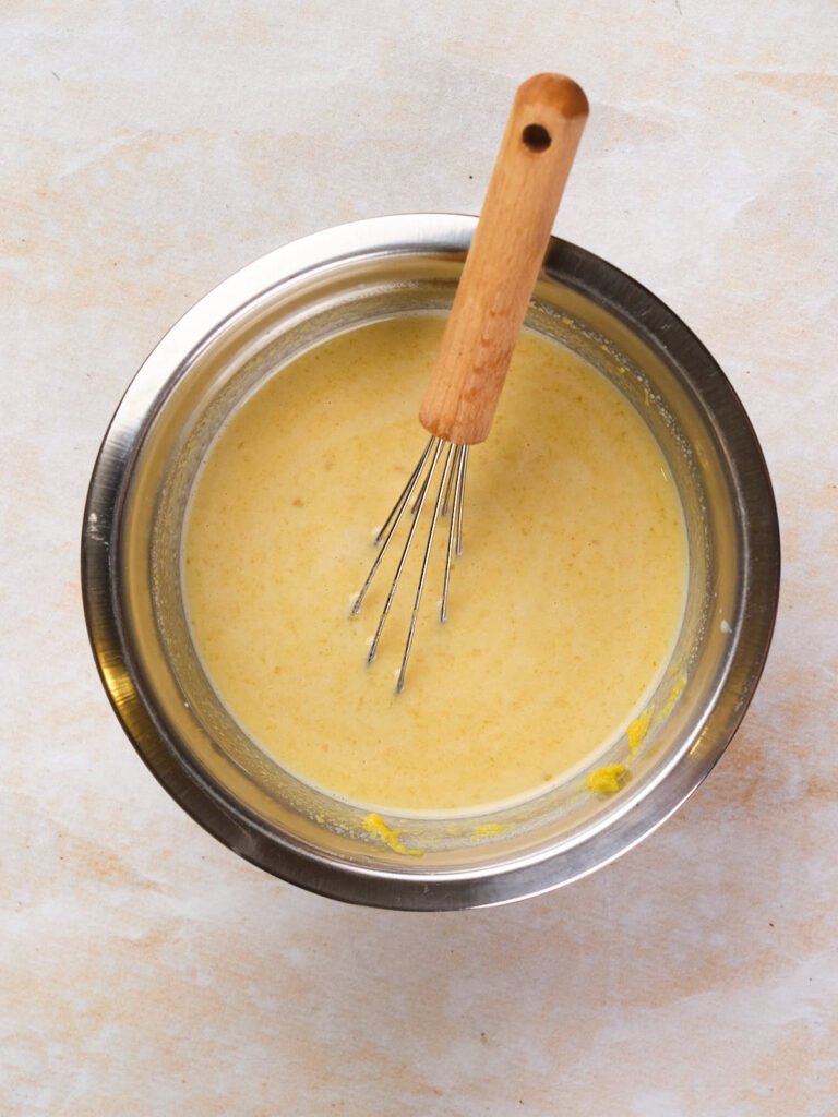 A metal mixing bowl filled with wet ingredients and a whisk with a wooden handle resting inside.