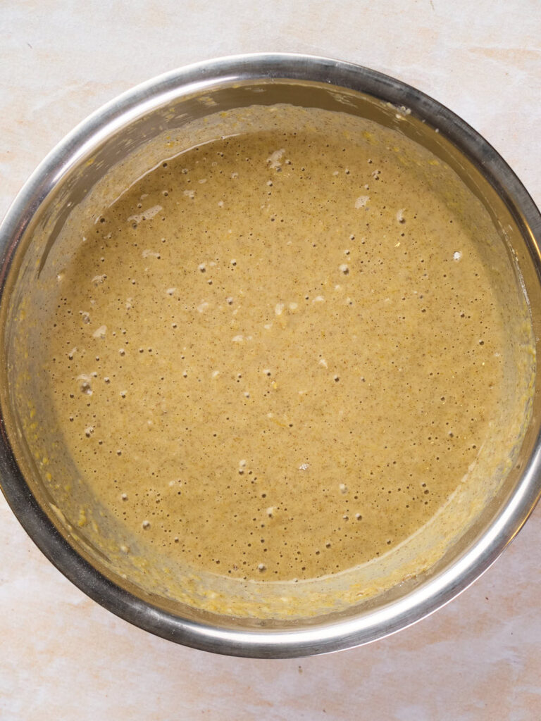 A metal mixing bowl filled with pancake batter dotted with small air bubbles, placed on a light-colored surface.