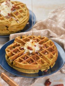 A stack of pumpkin pecan waffles is topped with butter and syrup being poured over them.