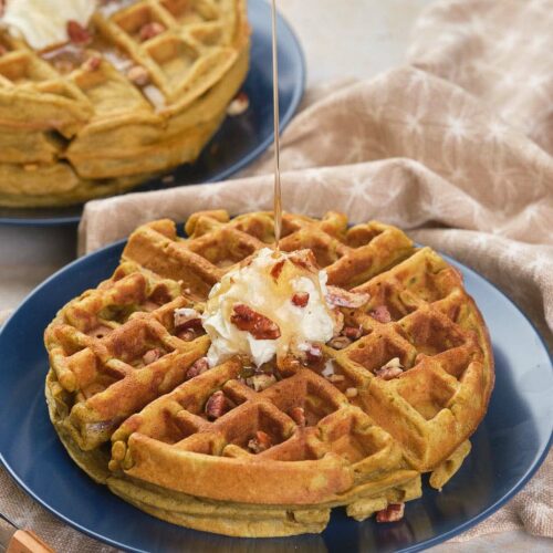 A stack of pumpkin pecan waffles is topped with butter and syrup being poured over them.