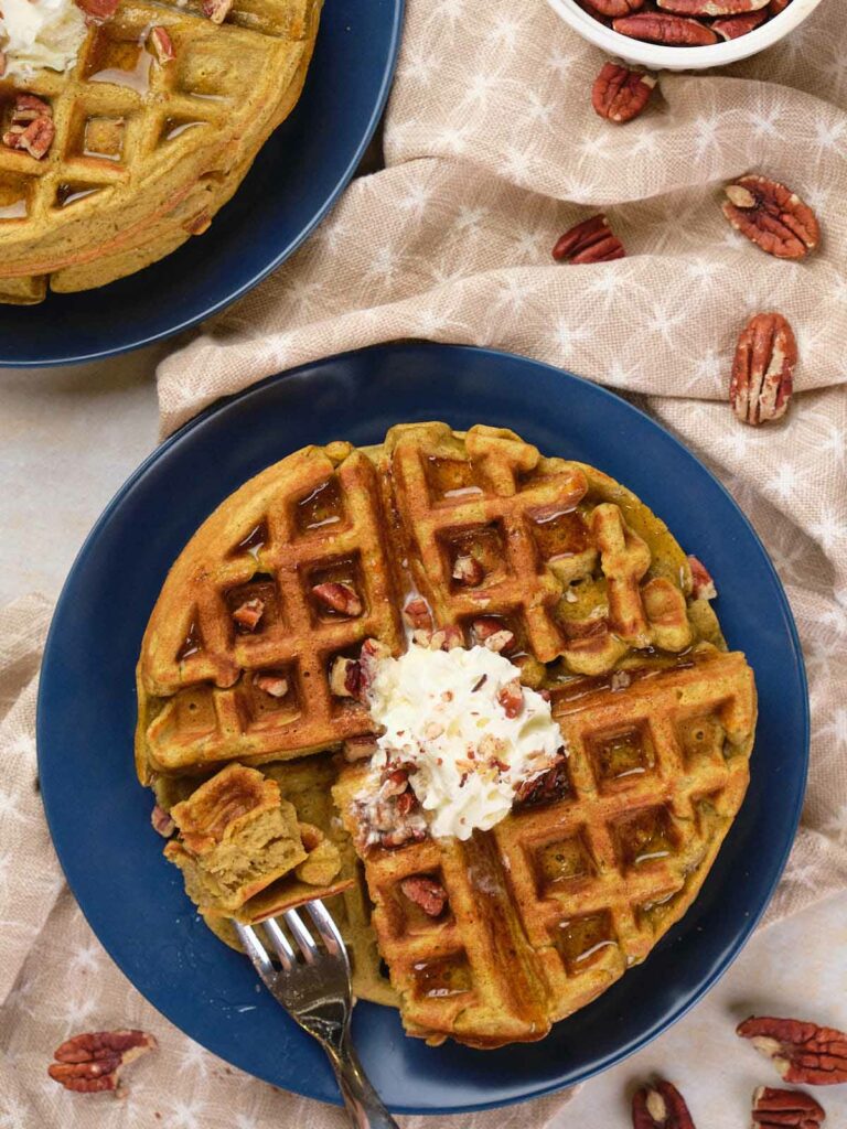 Two pumpkin pecan waffles on blue plates, topped with cream.