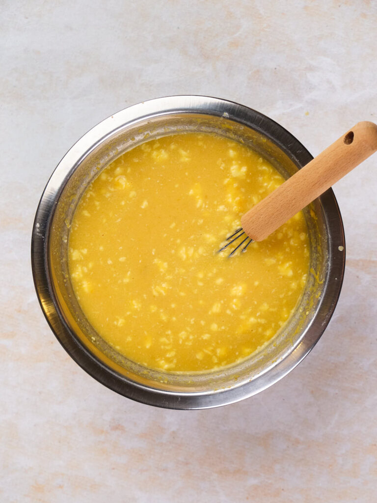 A metal bowl containing a yellow, lumpy mixture with a wooden-handled whisk resting on the side.