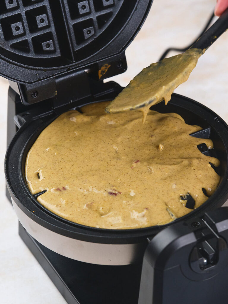 A waffle maker being filled with batter, using a spatula.