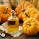 A small glass bottle filled with a golden liquid, surrounded by miniature pumpkins and pumpkin seeds.