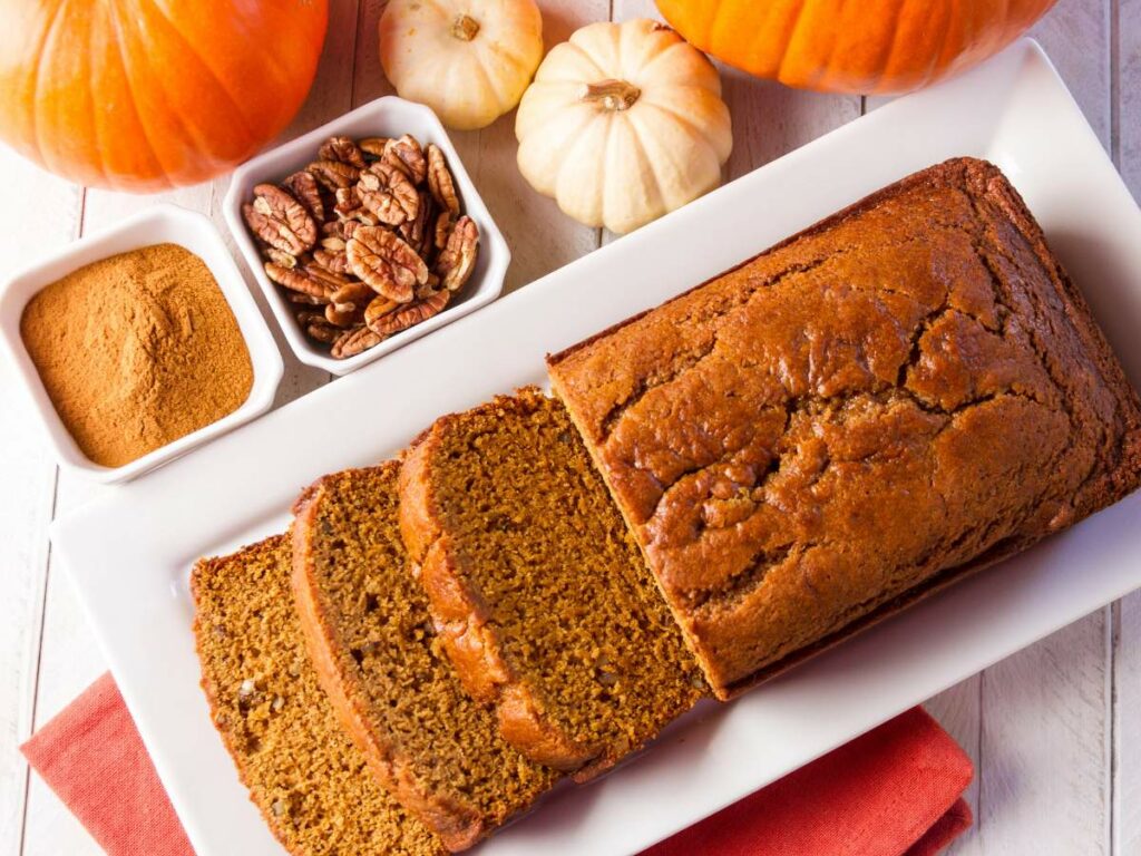 A rectangular white plate with sliced pumpkin bread, next to bowls of cinnamon powder and pecans.