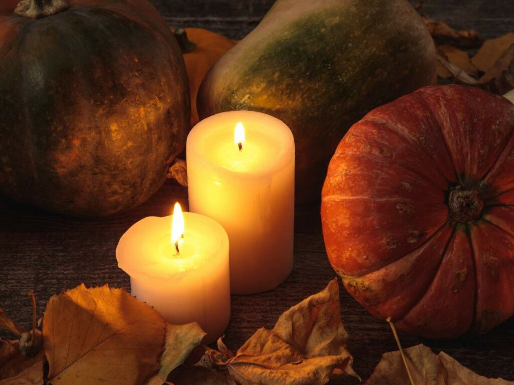 Two lit candles surrounded by pumpkins and fallen autumn leaves.