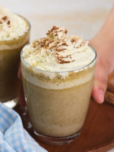 A hand holds a glass of pumpkin spice frappuccino topped with whipped cream.