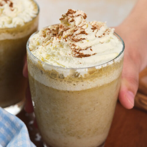 A hand holds a glass of pumpkin spice frappuccino topped with whipped cream.