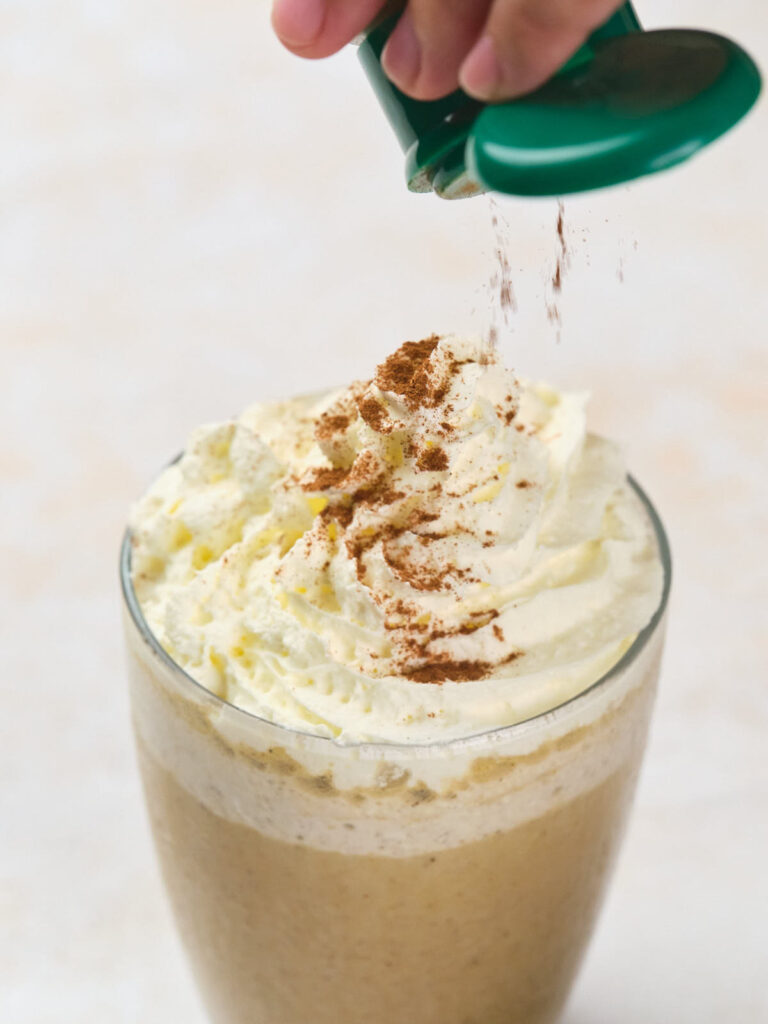 A hand sprinkles pumpkin spice over a whipped cream-topped frappuccino in a clear glass.