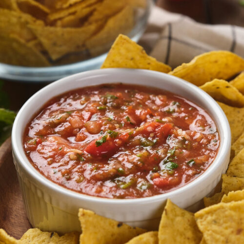 A bowl of salsa ranchera surrounded by tortilla chips on a wooden surface.