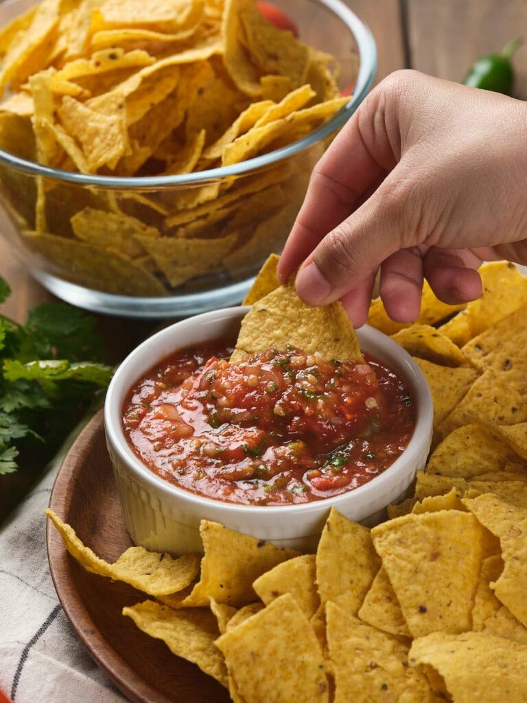 A hand dips a tortilla chip into a bowl of salsa ranchera , with a bowl of tortilla chips in the background.