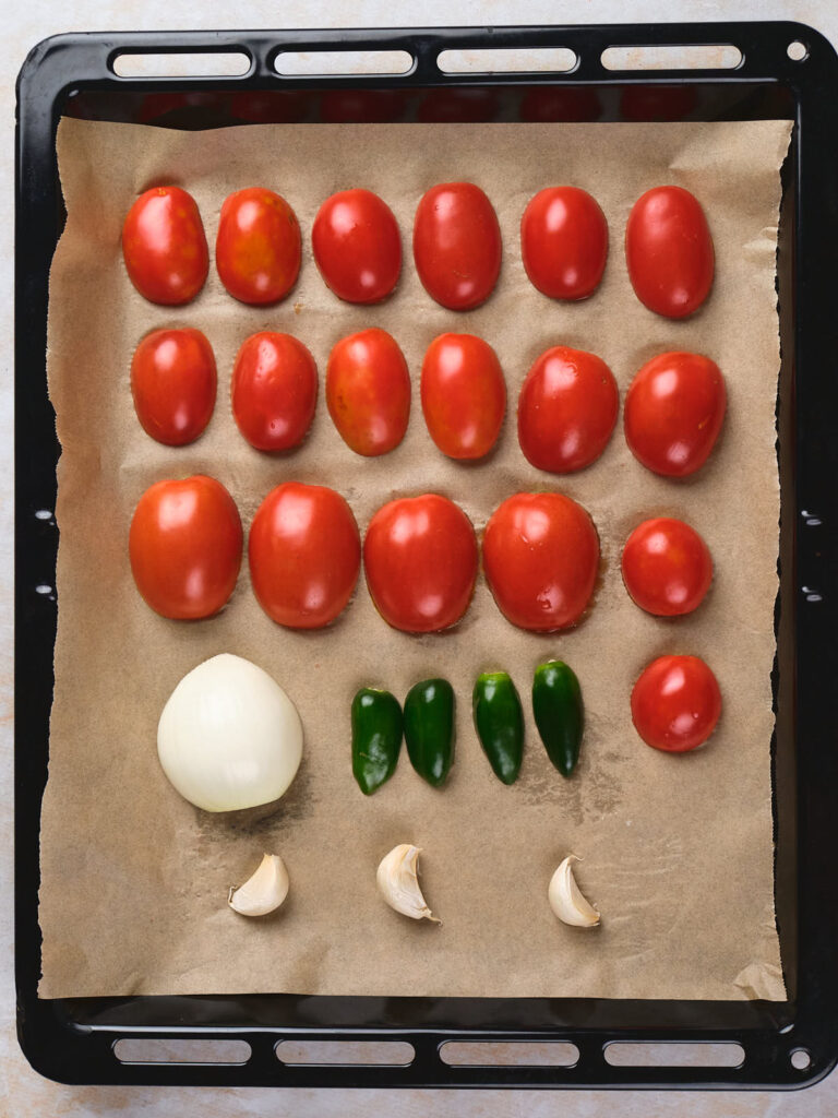 A baking tray lined with parchment paper holds rows of halved tomatoes, three whole jalapeños, a whole onion, and three garlic cloves.