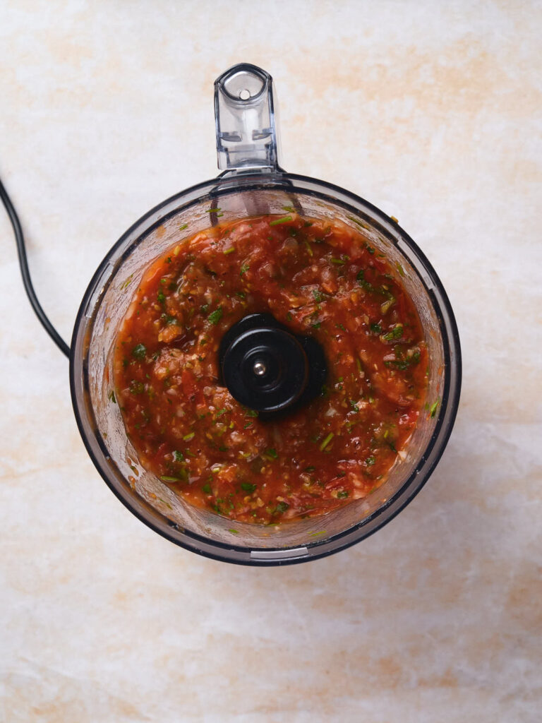 A glass bowl food processor containing salsa ranchera. The processor is viewed from above.