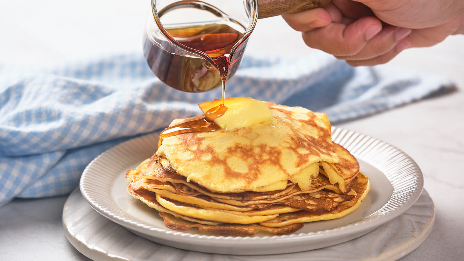 A person pours syrup over a stack of pancakes topped with butter on a white plate.