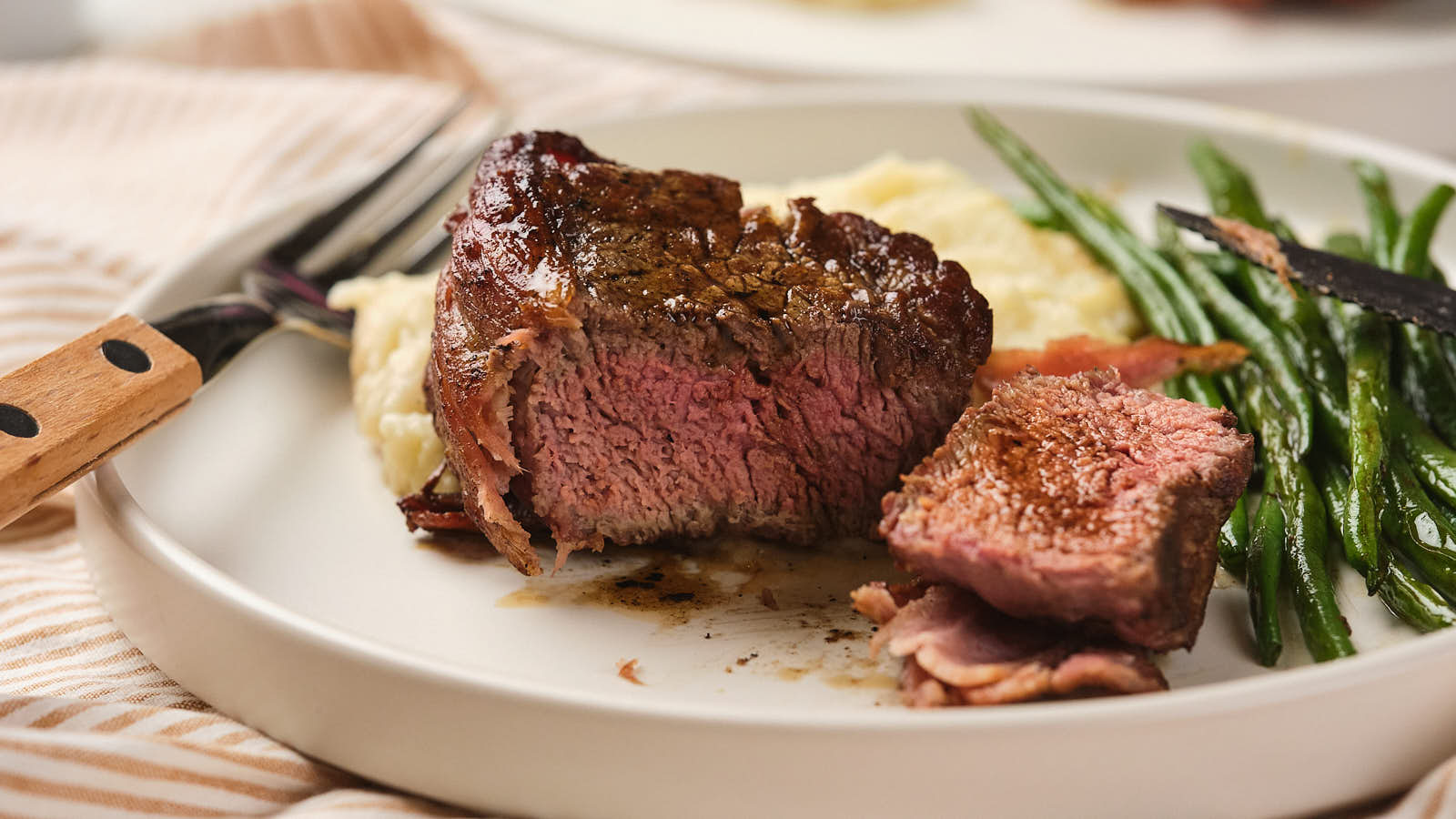 A plate with a cooked steak cut into pieces, accompanied by mashed potatoes and green beans.