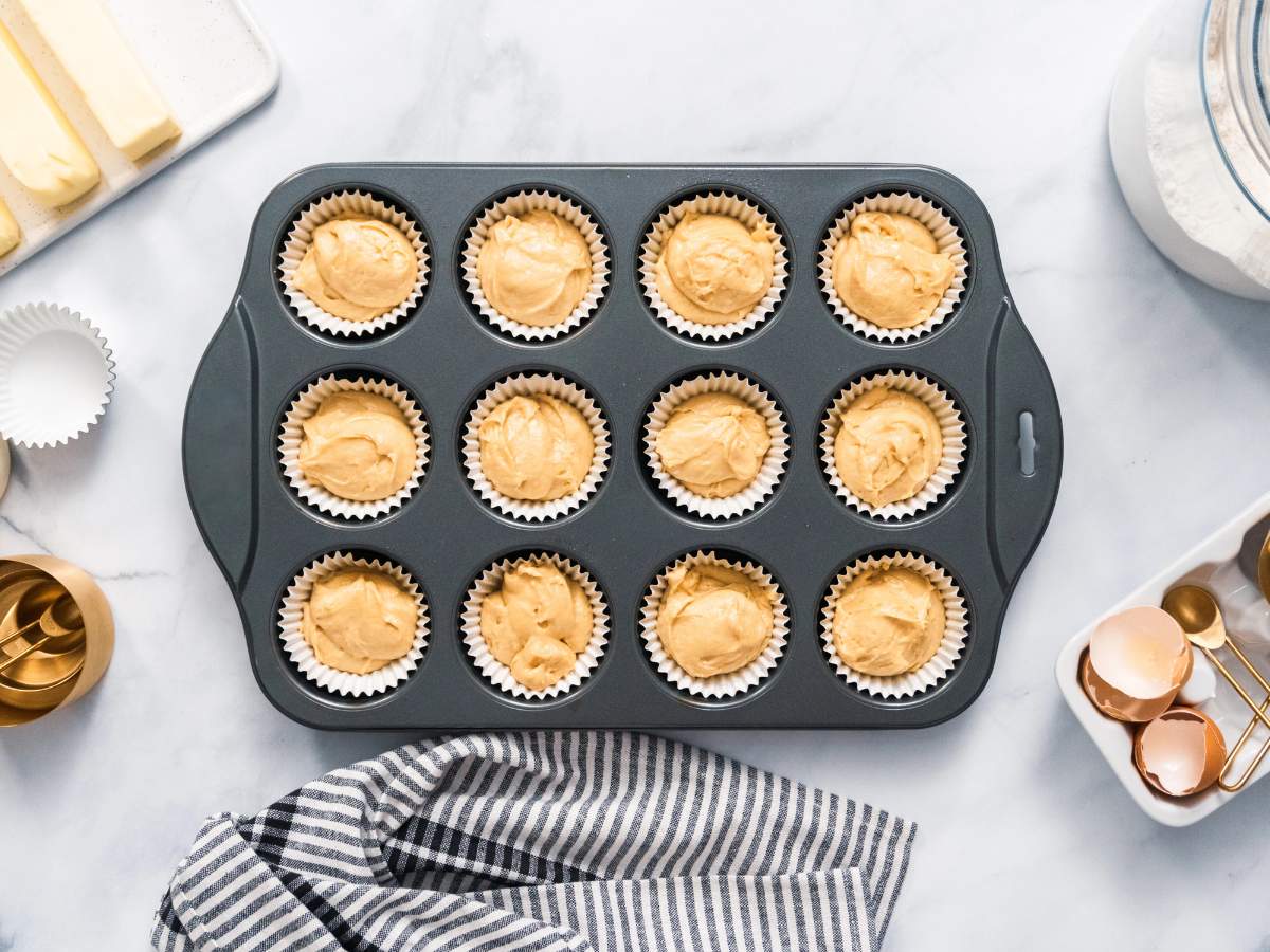 Cupcake batter in a 12-cup muffin pan surrounded by baking ingredients and tools on a marble countertop.