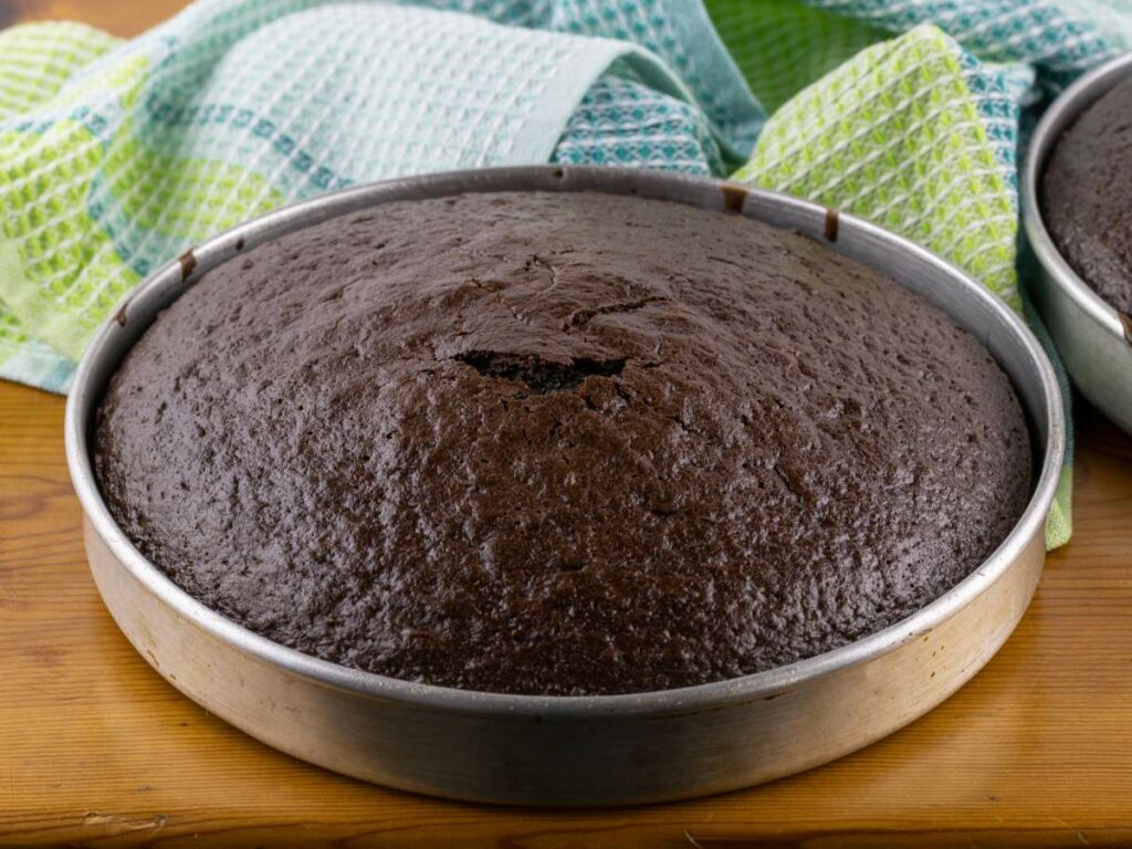 A freshly baked chocolate cake in a round metal pan on a wooden surface, with green and blue kitchen towels in the background.