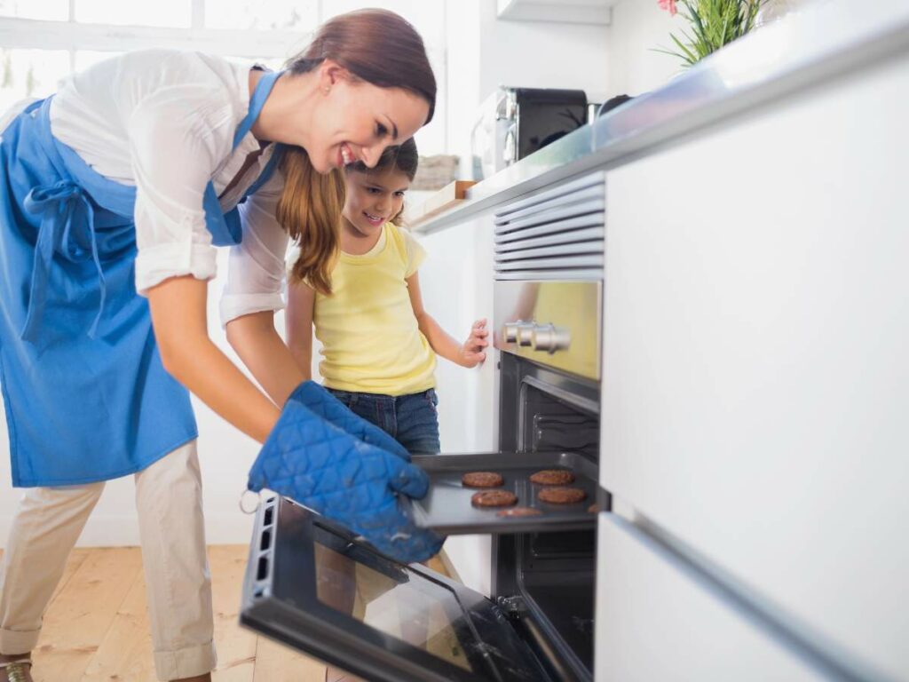 Woman in blue apron and oven mitts takes cookies out of the oven, while a young girl in a yellow shirt watches nearby in a bright kitchen.