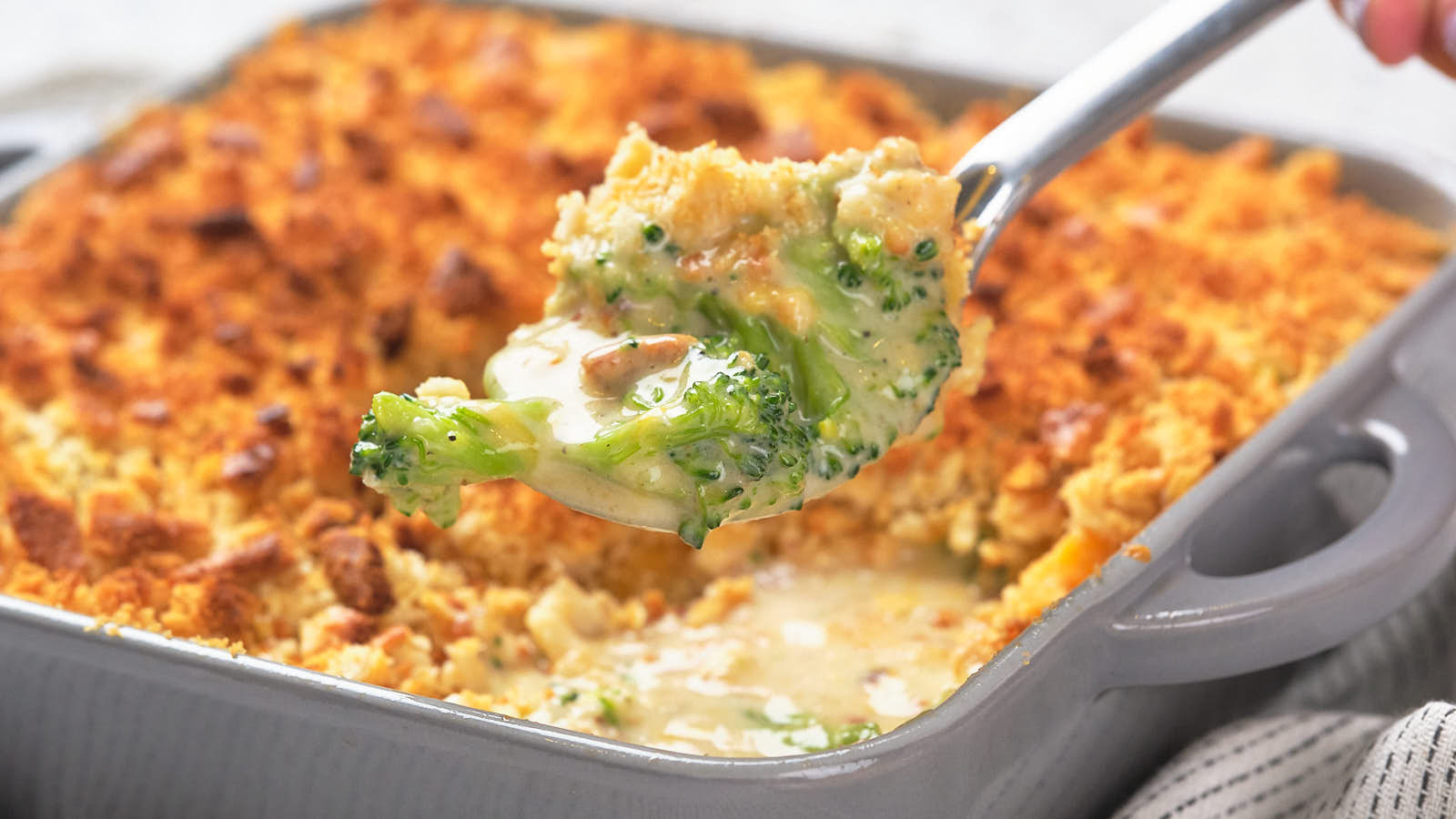 Close-up of a spoon scooping creamy broccoli casserole topped with a golden, crispy layer from a baking dish.